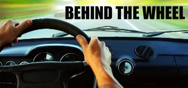 View out the front windshield of a car; text: Behind the Wheel